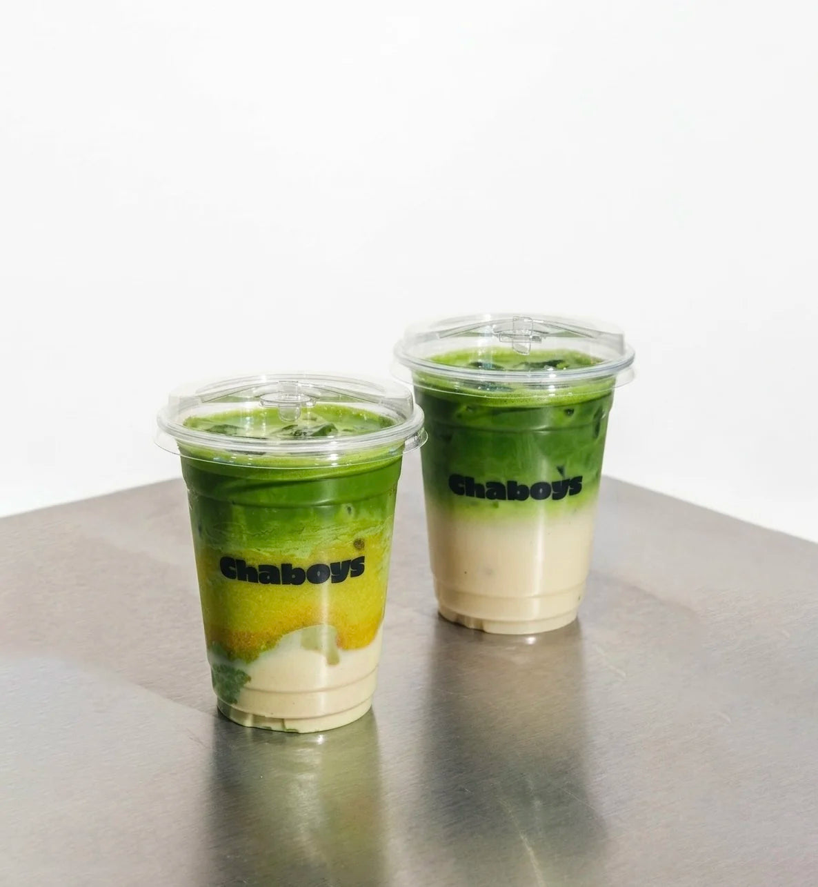 Two Chaboys matcha latte placed on a metallic gray surface against a white background.