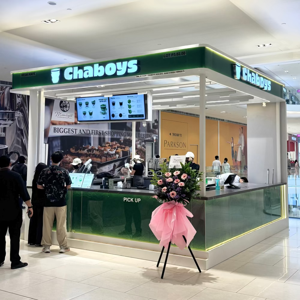 A Chaboys food kiosk in Pavilion Bukit Jalil shopping mall, featuring a green and white structure with digital screens, customers ordering, and a pink flower arrangement in the foreground.
