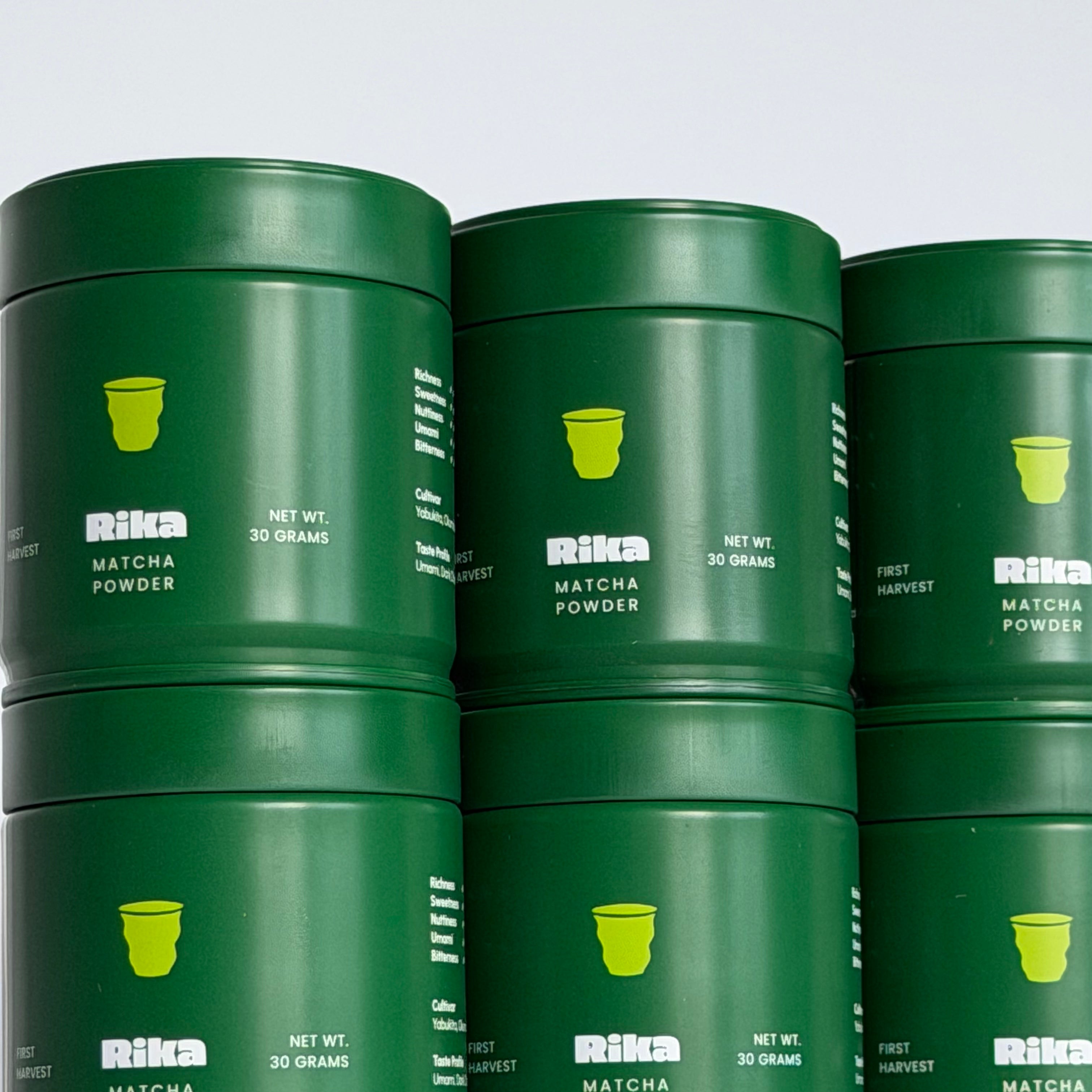 A stack of dark green cylindrical metal canisters labeled 'Rika Matcha Powder' with a small green Chaboys cup logo on each canister, photographed against a light gray studio background.